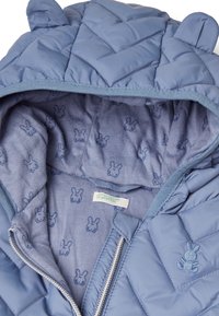 Padded modrá bunda s kapucí a ušima medvěda, obsahující prošívaný vnější povrch a podšívku s malými vzory králíčků. Stříbrný zip.