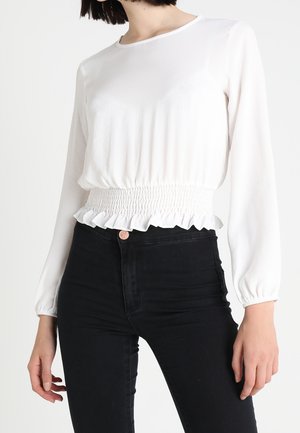 White long-sleeve top with a smocked waist and ruffled hem, paired with high-waisted black jeans. Smooth texture and relaxed fit.