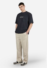 Tricou larg, negru, cu logo embosat, pantaloni bej cu croială largă și dungi pe laterale, adidași negri și albi. Texturi de țesătură netedă.