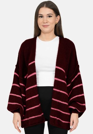 Femme portant un cardigan bordeaux ample avec des rayures horizontales roses par-dessus un haut blanc et un pantalon noir, debout devant un fond uni.
