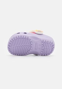 Suela de una pequeña zapatilla Crocs de color lavanda con un diseño de suela texturizada y marcas de tamaño, mostrando parte de la colorida correa superior.