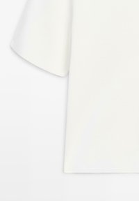 Chemise blanche à manches courtes avec un design simple, un ourlet droit et une texture de tissu douce, présentée sur un fond blanc.