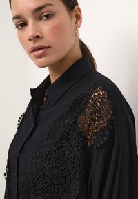 Maglietta nera con dettagli in pizzo intricati sulle spalle, tessuto morbido e colletto classico con accenti decorativi.