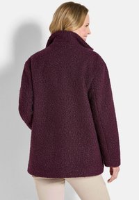 Plüschige, dunkelviolette Fleecejacke mit hohem Kragen, strukturiertem Finish, langen Ärmeln und lockerer Passform, von hinten betrachtet.