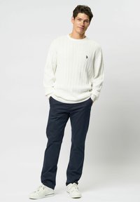 Tânăr stând cu mâinile în buzunare, purtând un pulover alb cu model torsadat, pantaloni închiși la culoare și adidași albi, pe un fundal luminos și simplu.