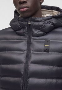 Gros plan d'une personne portant une doudoune noire avec capuche, fermeture éclair à l'avant et petit logo jaune Blauer sur la poitrine.