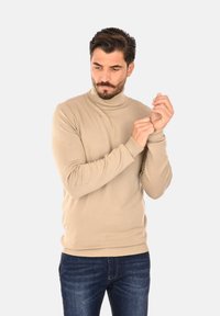 Maglione beige a collo alto lavorato a maglia con polsini e orlo a costine, abbinato a jeans in denim scuri. Texture liscia, design aderente.