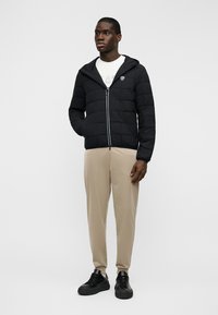 Μαύρο μπουφάν με επένδυση, με κουκούλα, φερμουάρ μπροστά και καπιτονέ σχέδιο, συνδυασμένο με μπεζ παντελόνι και μαύρα αθλητικά.