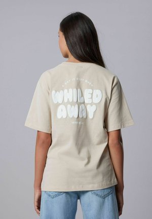 Mujer con cabello largo y oscuro que lleva una camiseta beige con el texto "Un día es mejor cuando se pasa de manera relajada versión 2.0", combinada con jeans de color azul claro.