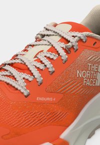 Κοντινό πλάνο λαμπερού πορτοκαλί αθλητικού παπουτσιού The North Face Enduris 4 με υφασμάτινη υφή και λευκά κορδόνια με σχέδιο σε λευκή σόλα.