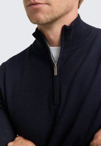 Mann in einem navyblauen Strickpullover mit halben Reißverschluss und geripptem Kragen über einem weißen Hemd, mit verschränkten Armen.