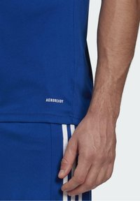 Niebieska koszulka sportowa z miękkiego materiału, z małym białym logo "AEROREADY" na dolnym brzegu, w połączeniu z pasującymi szortami i paskowymi akcentami.