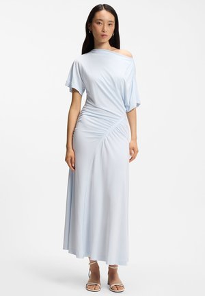 Frau trägt ein hellblaues, schulterfreies, gerafftes Maxikleid mit kurzen Ärmeln und weißen Knöchelriemen-Absätzen, steht vor einem weißen Hintergrund.