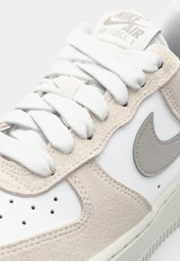 Nike Air Force 1 sneakers med en overdel i hvid læder, beige ruskindsdetaljer og flade hvide snørebånd. Designet inkluderer en grå Nike swoosh.