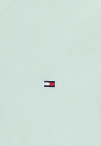 Petit logo rectangulaire brodé avec des sections noires, blanches et rouges sur un tissu texturé vert clair.