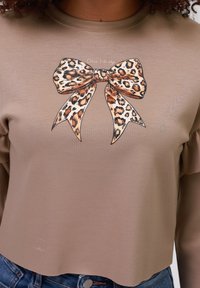 Blusa beige de manga larga con un diseño recortado que presenta un gran gráfico de un lazo de leopardo en el centro y un sutil detalle de texto por encima.