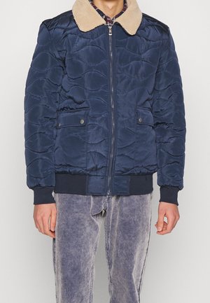 Homme portant une veste matelassée bleu marine avec un col en laine beige et des poches avant, assortie à un pantalon en velours côtelé gris clair.