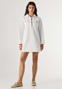 Abito in denim bianco con colletto, maniche lunghe e due tasche sul petto. Presenta una patta di bottoni e una sagoma leggermente svasata.