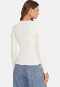 Top bianco a maniche lunghe a coste con design aderente e scollo rotondo, indossato con jeans a vita alta azzurri.