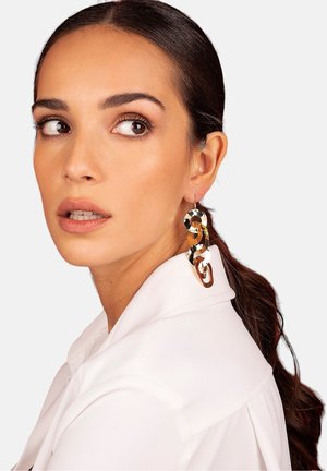 Gold and silver twisted hoop earrings with a glossy finish, featuring a sculptural design. The model wears a white collared shirt.