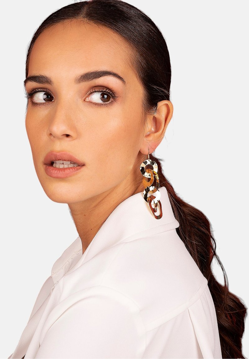 Orecchini a cerchio attorcigliati in oro e argento con una finitura lucida, caratterizzati da un design scultoreo. Il modello indossa una camicia bianca con colletto.