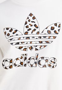 Hvit bomulls-T-skjorte med leopardmønstret Adidas-logo. Har tre bladlignende former over ordet "adidas," med svarte og brune detaljer.