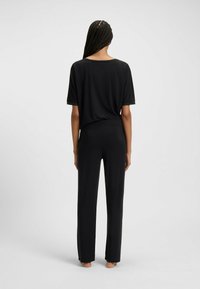 Vrouw met gevlochten haar die blootsvoets staat, gekleed in een losse zwarte top met korte mouwen en zwarte wijde broek, met de rug naar de camera gekeerd tegen een eenvoudige achtergrond.