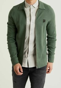 Grön zip-up jacka med krage, ribbad textur och en liten logotypapplikation på vänster bröst. Bärs över en ljus knapp-up skjorta.