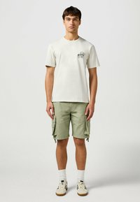 Hellbeige T-Shirt mit einem kleinen Grafikmotiv, kombiniert mit olivgrünen Cargo-Shorts mit Seitentaschen. Weiße Socken und cremefarbene Sneaker vervollständigen das Outfit.
