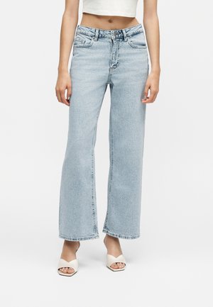 Kvinde iført lyseblå jeans med brede ben, hvid croppet top og hvide åbne hælede sandaler, stående mod en ensartet baggrund.