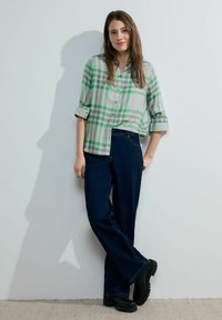 Camicia button-up a quadri verde e grigio con maniche arrotolate, abbinata a jeans a gamba larga blu scuro e stivali platform neri.