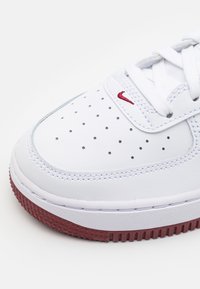 White leather sneaker with perforated toe box, red Nike swoosh, and textured red rubber outsole. Laces are white, stitching is visible.