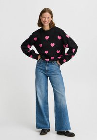 Maglione nero con motivi a cuore rosa, abbinato a jeans blu a vita alta e gamba larga e scarpe nere. Look informale, vestibilità e design rilassati.
