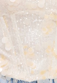 Gros plan d'un tissu beige avec de grands sequins ronds et de petites perles brillantes sur un fond en denim bleu clair.