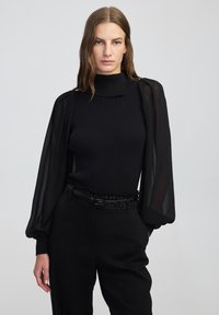 Top lungo a maniche ribbed nere con maniche puff trasparenti, caratterizzato da un collo alto e una cintura nera texturizzata con decorazioni.