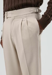 Beige op maat gemaakte broek met scherpe plooien, zijzakken en een riem met een metalen gesp, gecombineerd met een geribbelde bruine trui.