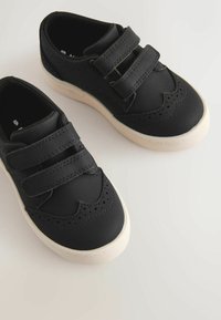 Melnas sneakers ar divām Velcro siksnām, gludu sintētisku augšdaļu, dekoratīvām perforācijām un baltu gumijas zolīti. Minimālistisks dizains.