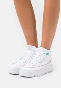 Białe trampki typu high-top z gładkiej skóry, różowymi i turkusowymi akcentami oraz grubą podeszwą. Posiadają logo na języku i boku.