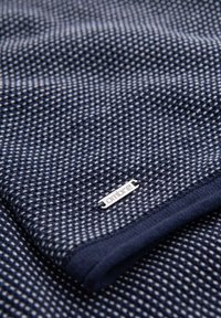 Navyblått stickat tyg med ett strukturerat mönster och en silverfärgad "ombre"-etikett på kanten. Mjukt, vävt material med en slät yta.