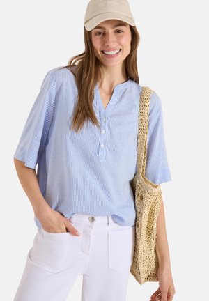 Femme souriante portant une casquette beige « OPTIMISME », un chemisier rayé bleu clair, un pantalon blanc, et portant un sac cabas tissé beige sur l'épaule.