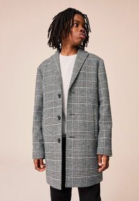 Cappotto a scacchi grigio e nero con rever, chiusura a bottoni e due tasche. Indossato sopra una camicia grigia chiara, esposto su uno sfondo neutro.