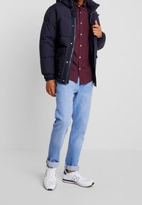 Mann in einer marineblauen Steppjacke über einem rot karierten Hemd, hellblauen hochgekrempelten Jeans und weißen New Balance Sneakers.