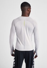 Maglia sportiva a maniche lunghe di colore grigio chiaro. Presenta un tessuto strutturato, un design aderente e un dettaglio riflettente giallo lungo la colonna vertebrale.