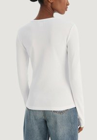 Bílé žebrované tričko s dlouhým rukávem s priléhavým střihem a kulatým výstřihem, v kombinaci s modrými džínami, zobrazené zezadu.