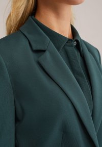 Mėlynas švarkas su aštriu apykakle ir glotnia tekstūra, dėvimas ant tamsiai žalių marškinių su mygtukų apykakle ir subtiliu audinio apdaila.