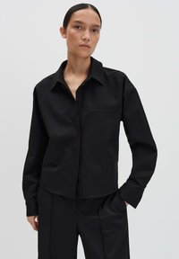 Vrouw met donker haar in een lage knot, gekleed in een zwart overhemd met kraag en zwarte broek, tegen een simpele lichte achtergrond.