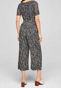 Svart kortärmad jumpsuit med halvgenomskinlig textur, prickigt mönster i kräm och grönt, inbunden midja, lös passform och vida ben.