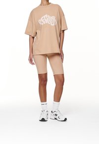 Person som bär en beige oversized t-shirt med texten "PEGDOR", matchande beige cykelbyxor, vita strumpor och vita sneakers från New Balance.