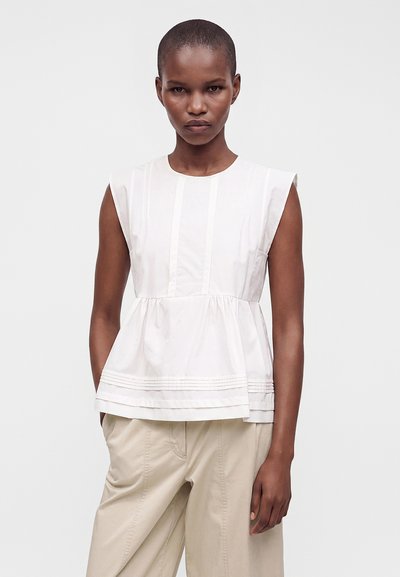 Femme aux cheveux courts portant un top blanc sans manches à basque et un pantalon beige, debout devant un fond clair uni.