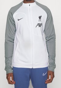 Chaqueta blanca con cremallera y mangas grises, con el logo del Liverpool FC, marca Nike y logo de AXA, fabricada en material sintético suave.
